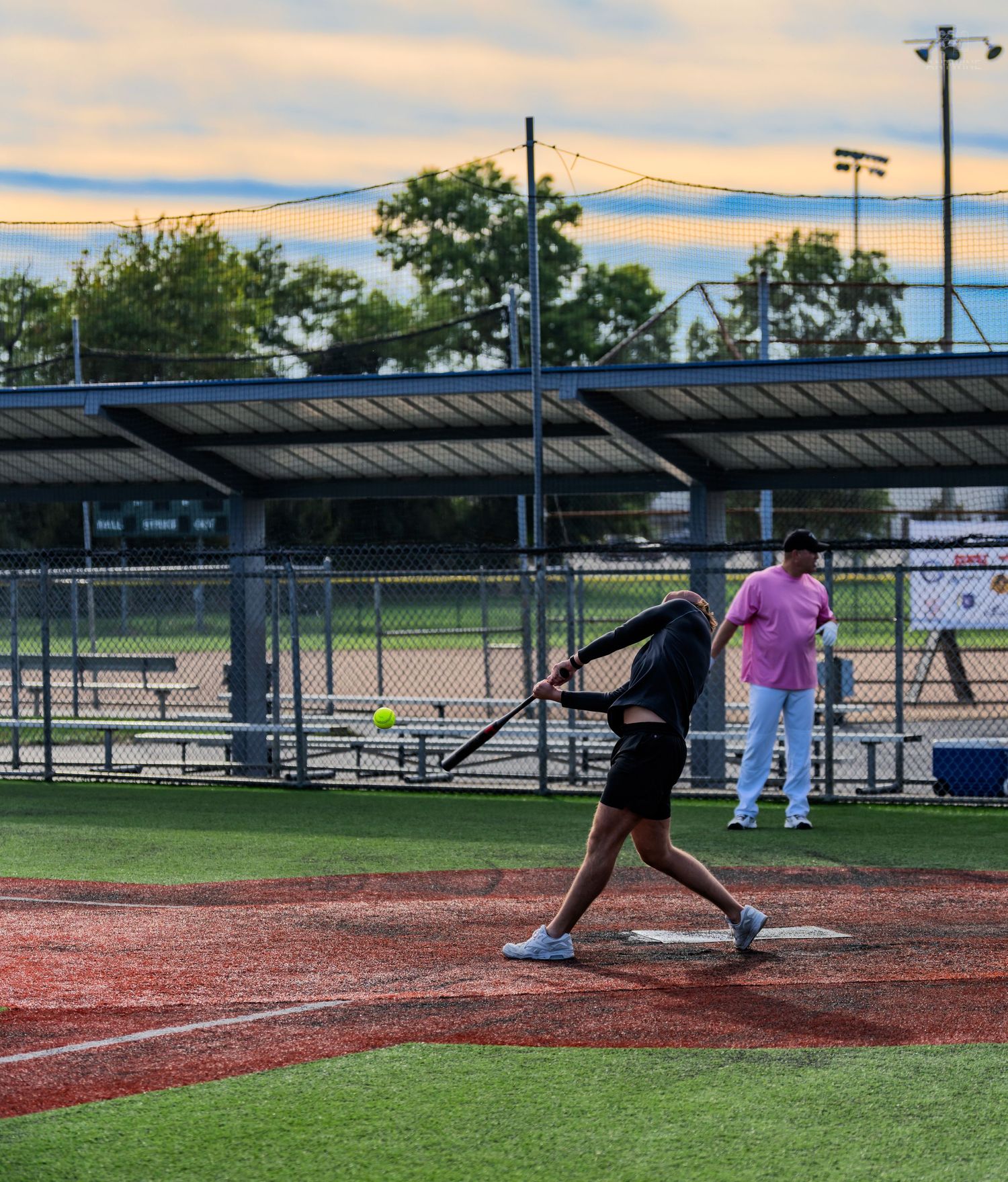 Player in black swings at a yellow softball during an evening recreational game at a well-lit baseball diamond.