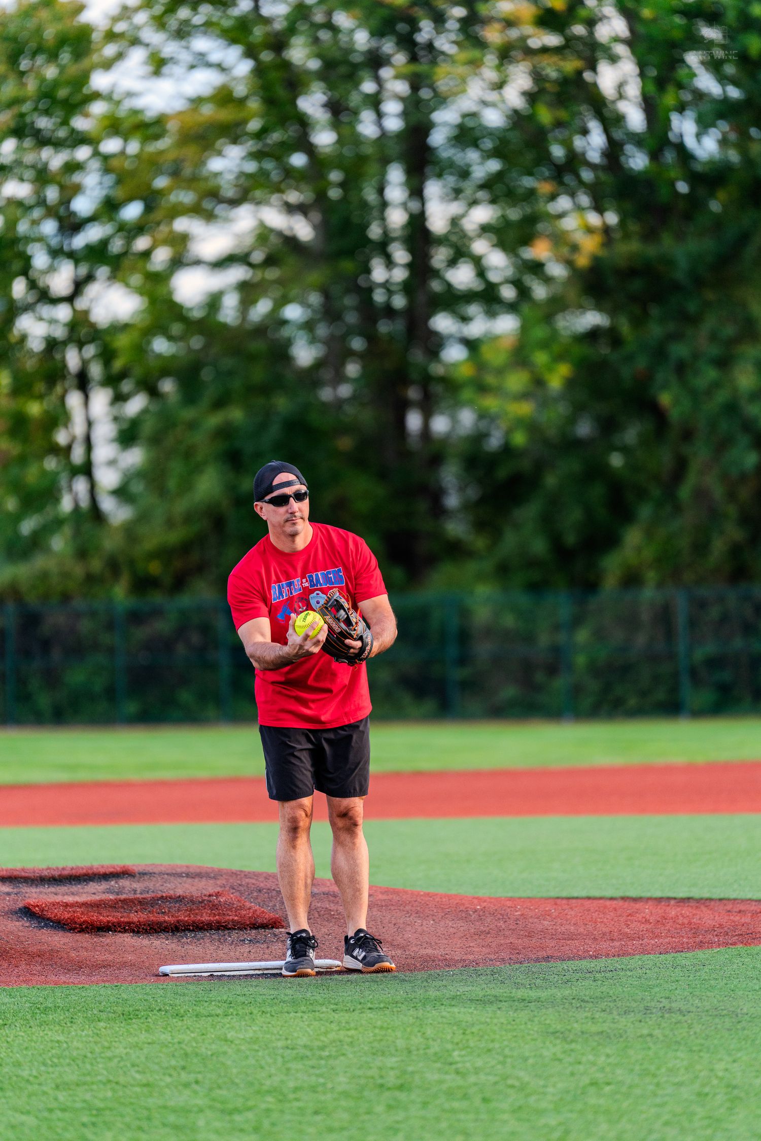 Man in red shirt pitching a yellow softball on a well-maintained baseball field outdoors.