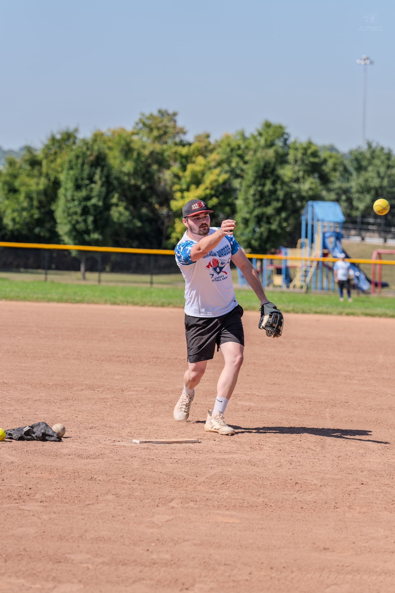 Man in white jersey throwing a softball on a dirt infield during an outdoor recreational softball game on a sunny day.