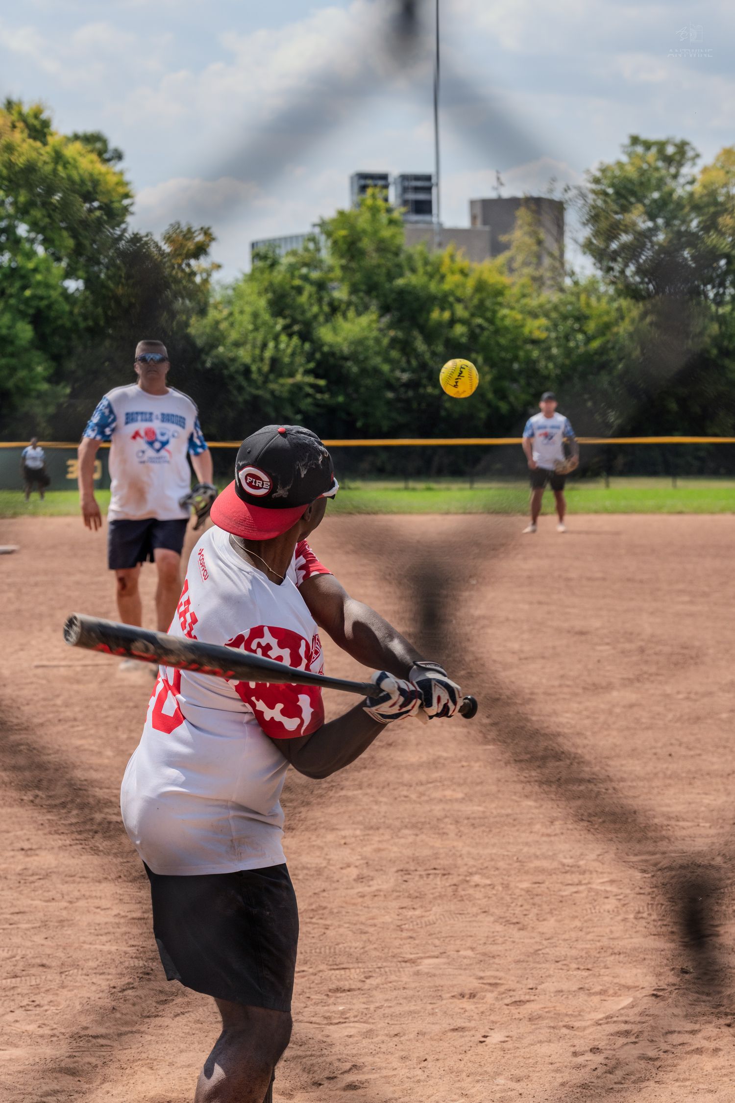A softball batter in a red cap swings at a yellow ball during an outdoor game on a dirt field.