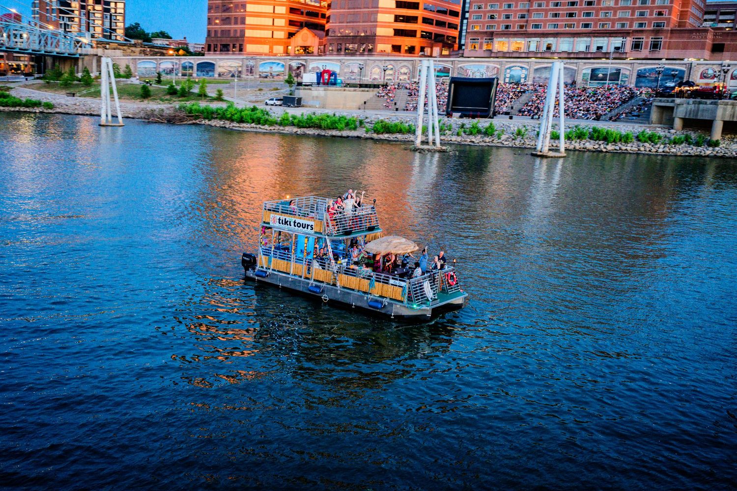 Aerial view of a colorful pontoon boat cruising on a river with urban buildings in the background.