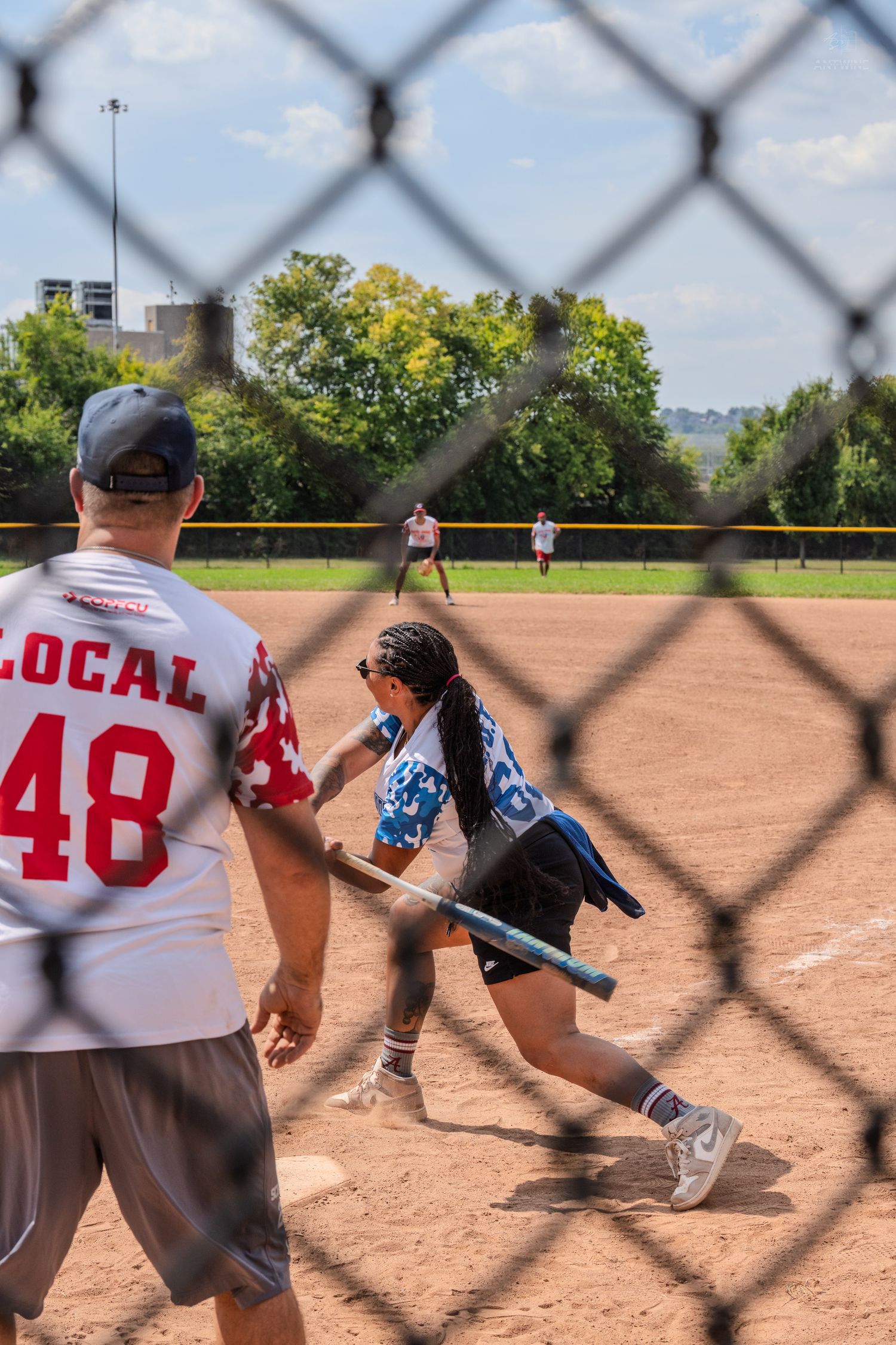 A batter swings during a charity softball game viewed through a chain-link fence with a coach in a Local 48 jersey nearby.