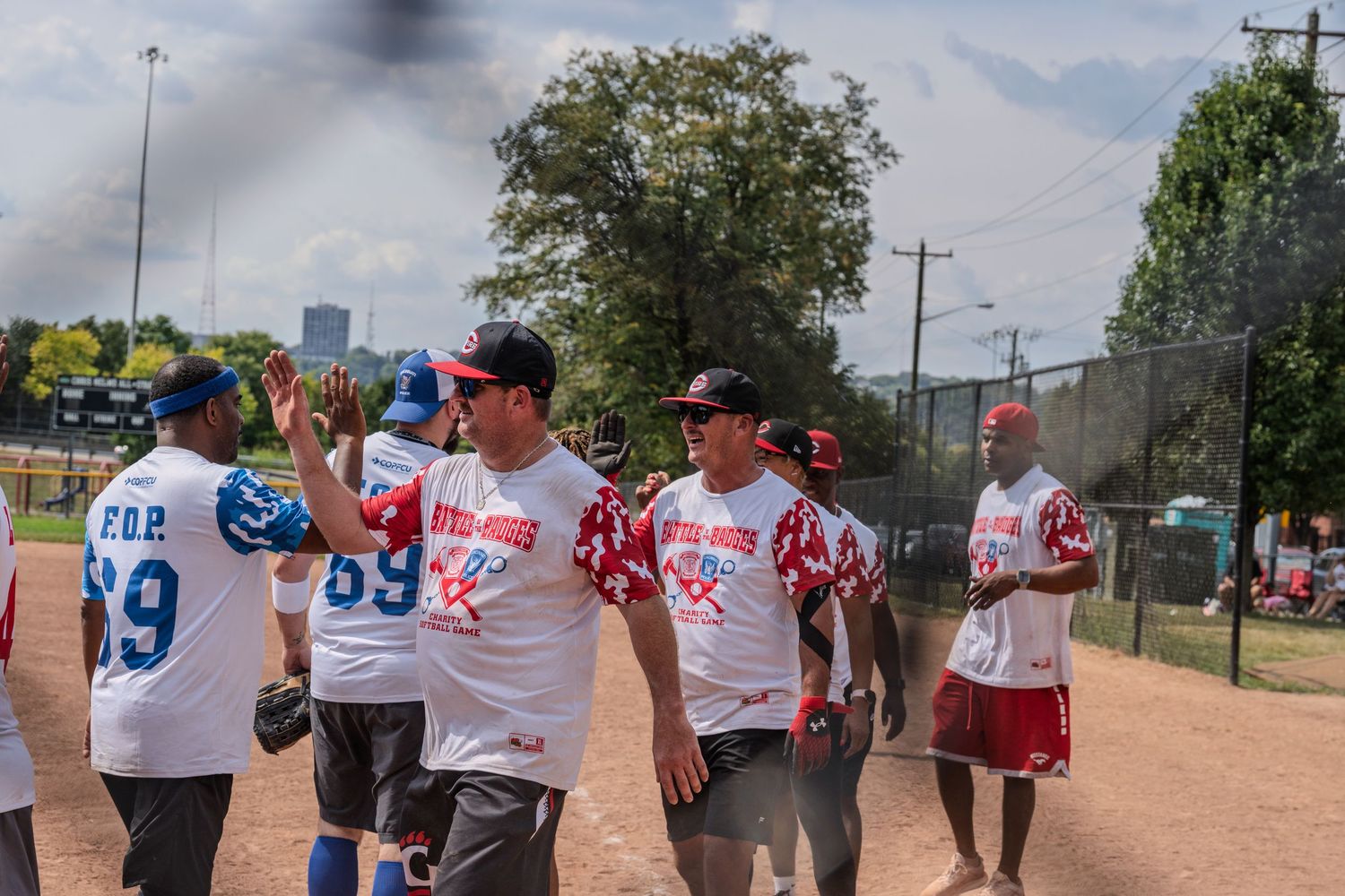 Softball players in red and white jerseys high-fiving on a dirt field during a sunny outdoor game.