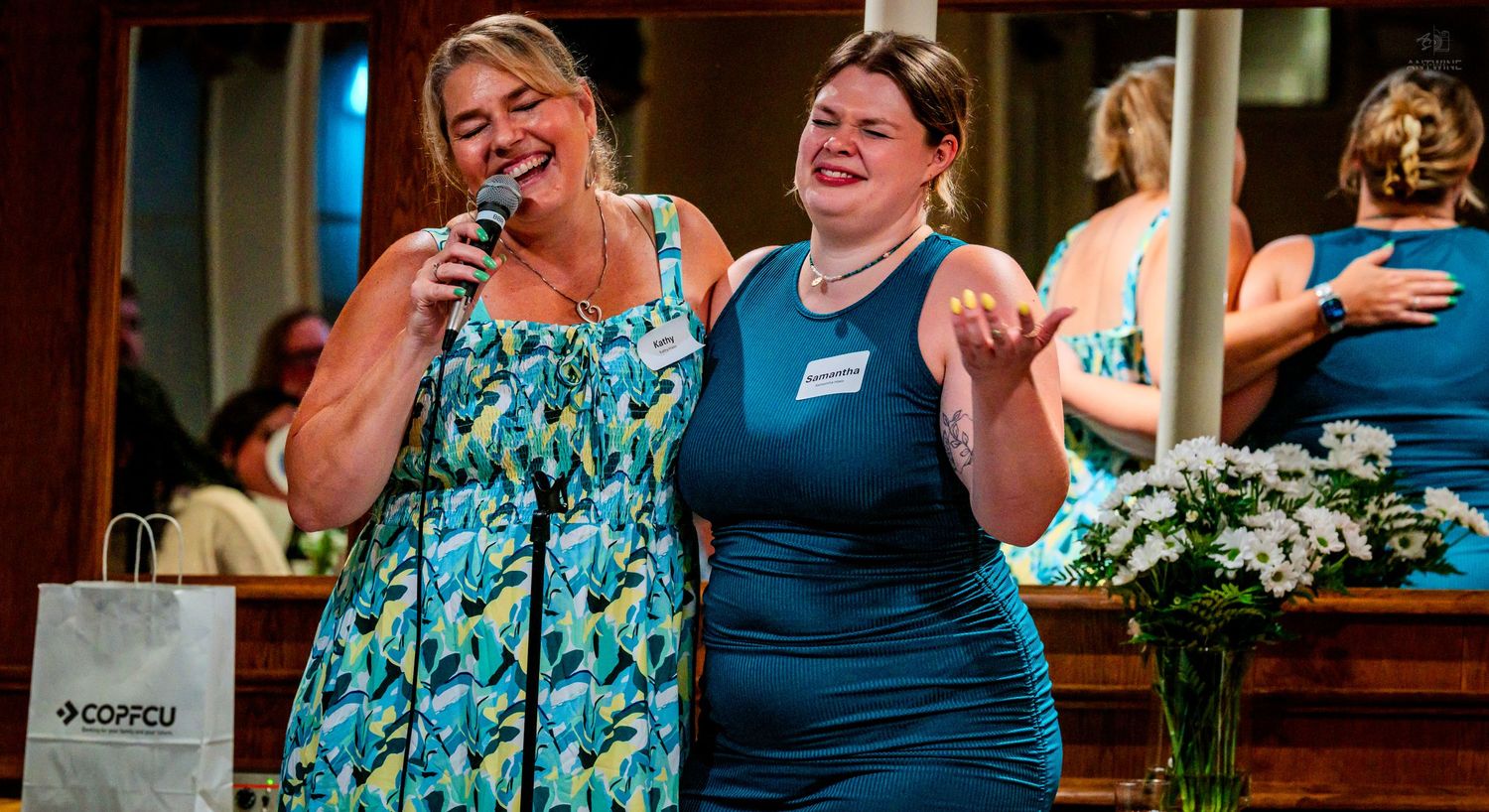 Two cheerful women stand together on stage at an indoor event with floral decor.