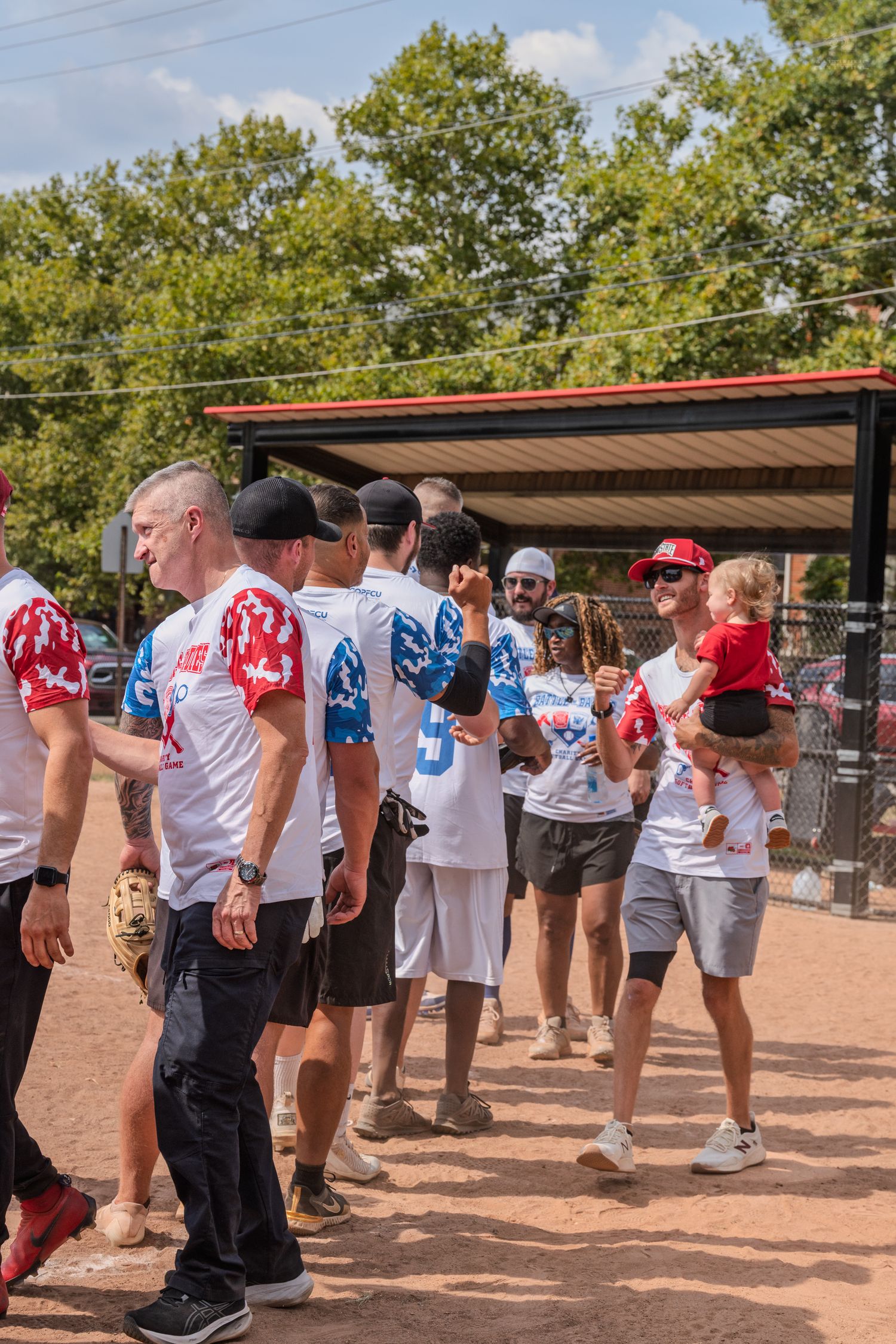 Softball players in red and white uniforms shaking hands on a baseball field after a charity game outdoors.