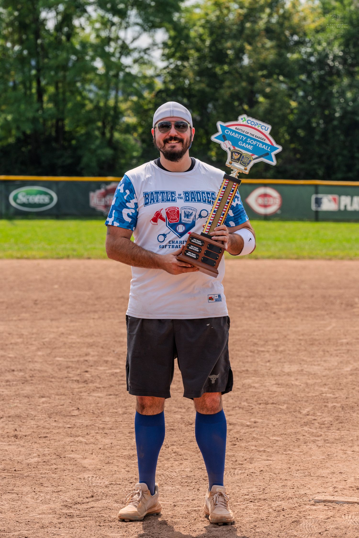 Man holds charity softball game trophy on baseball field wearing Battle of the Badges shirt.