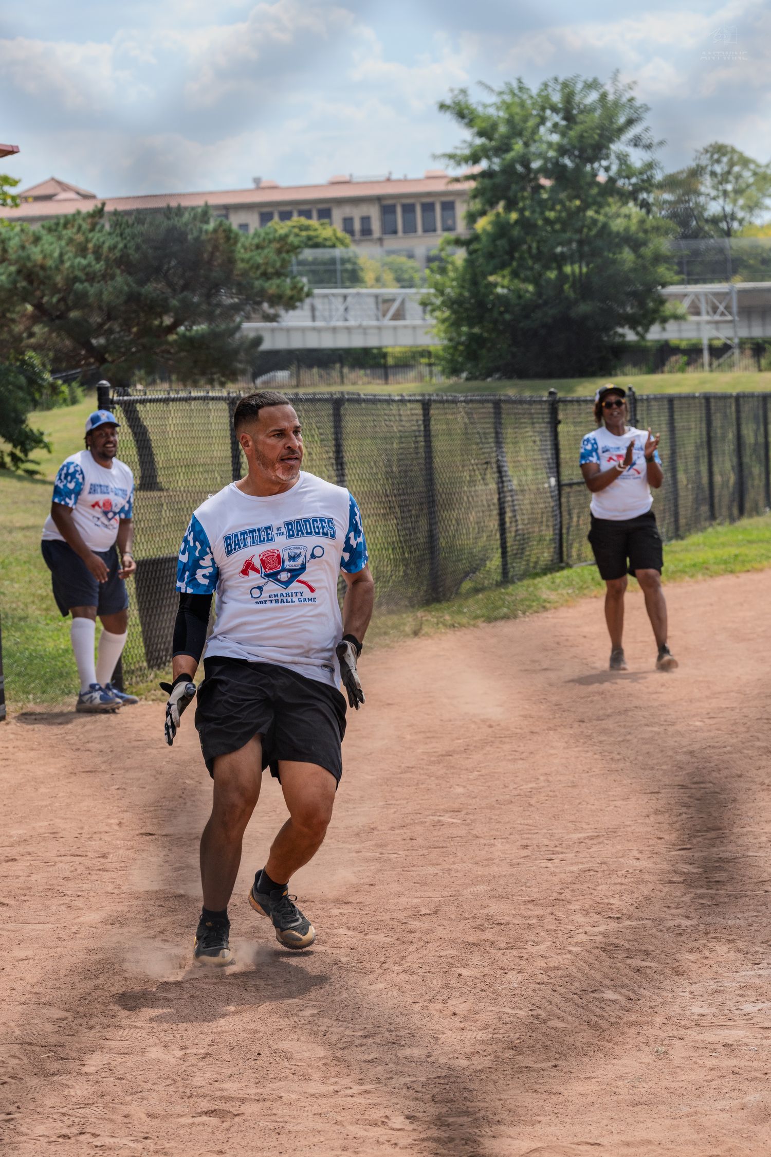 Softball player in charity game jersey jogs along baseline at outdoor baseball field.