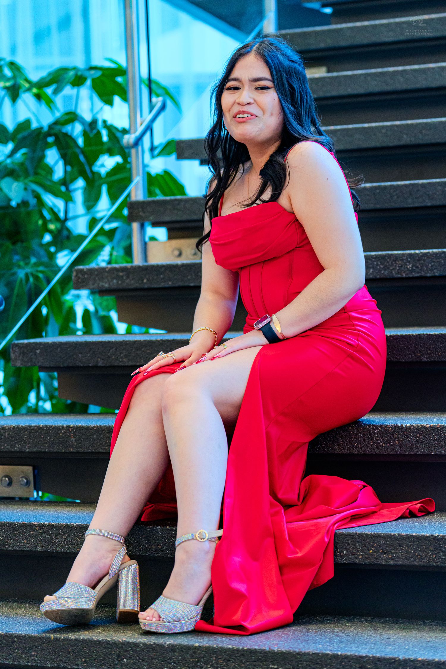 Young woman in red off-shoulder formal dress sitting on outdoor stairs smiling at camera.