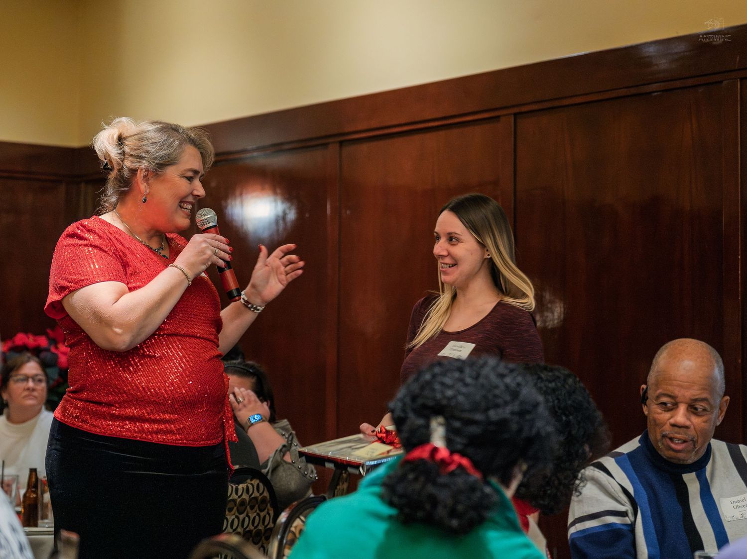 Woman in red speaks into microphone at indoor event while another woman smiles beside her at a banquet hall.