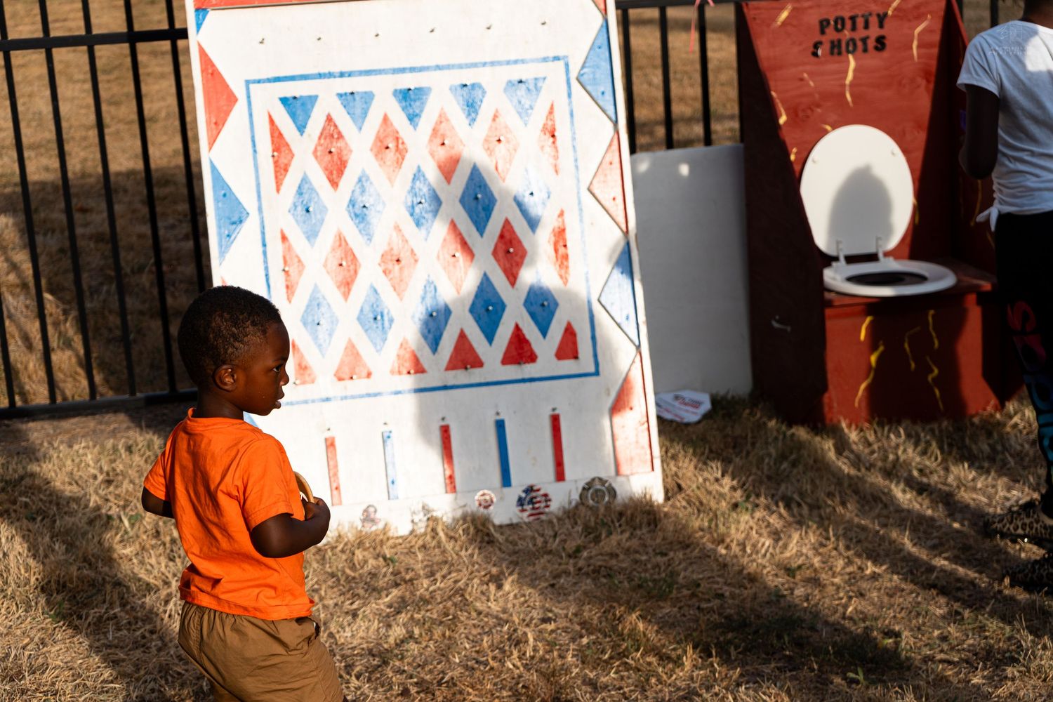 Young Black child in orange shirt standing near colorful diamond-patterned game board at outdoor carnival.