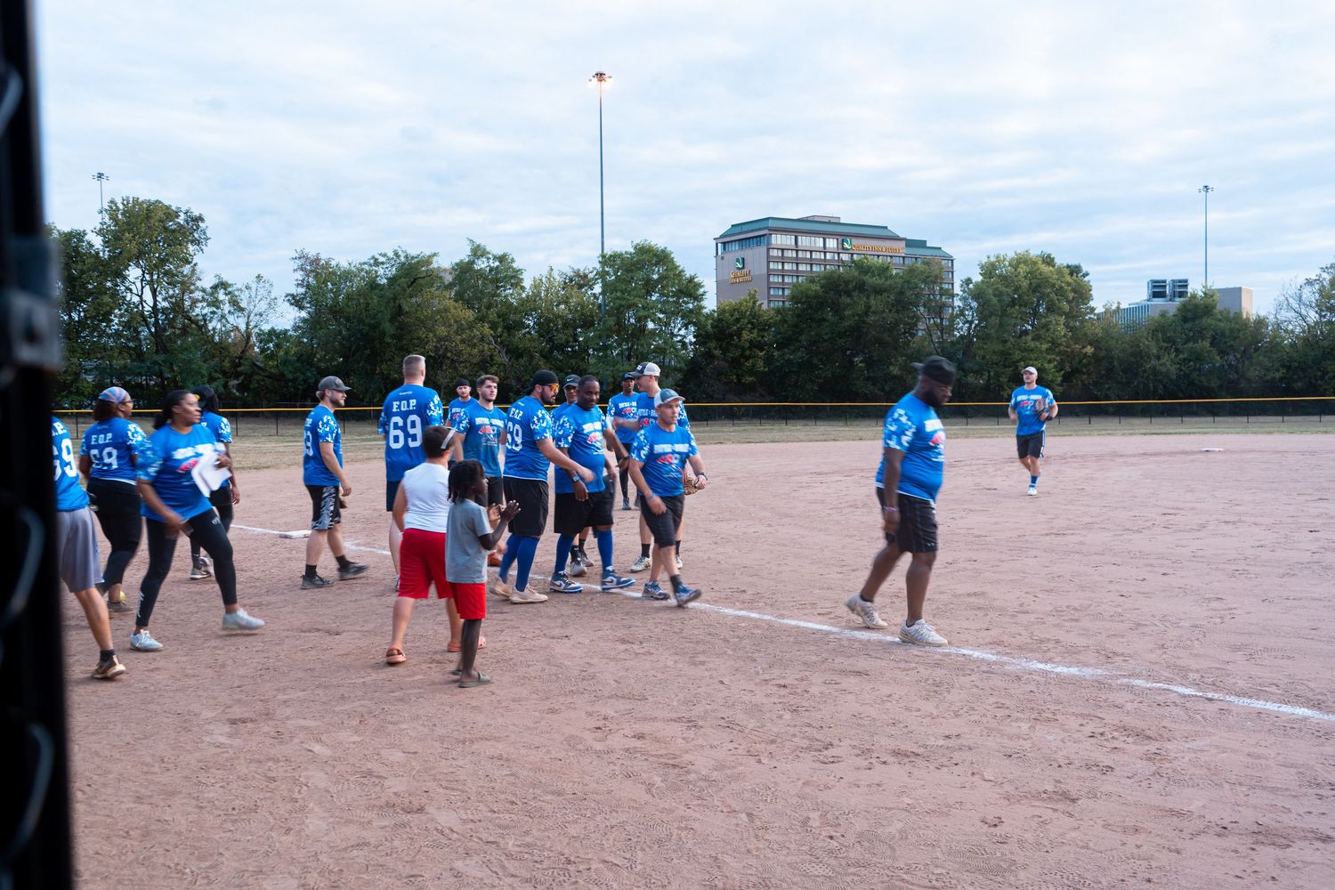 Group of softball players in blue uniforms gathered on a dirt field during an outdoor community game.