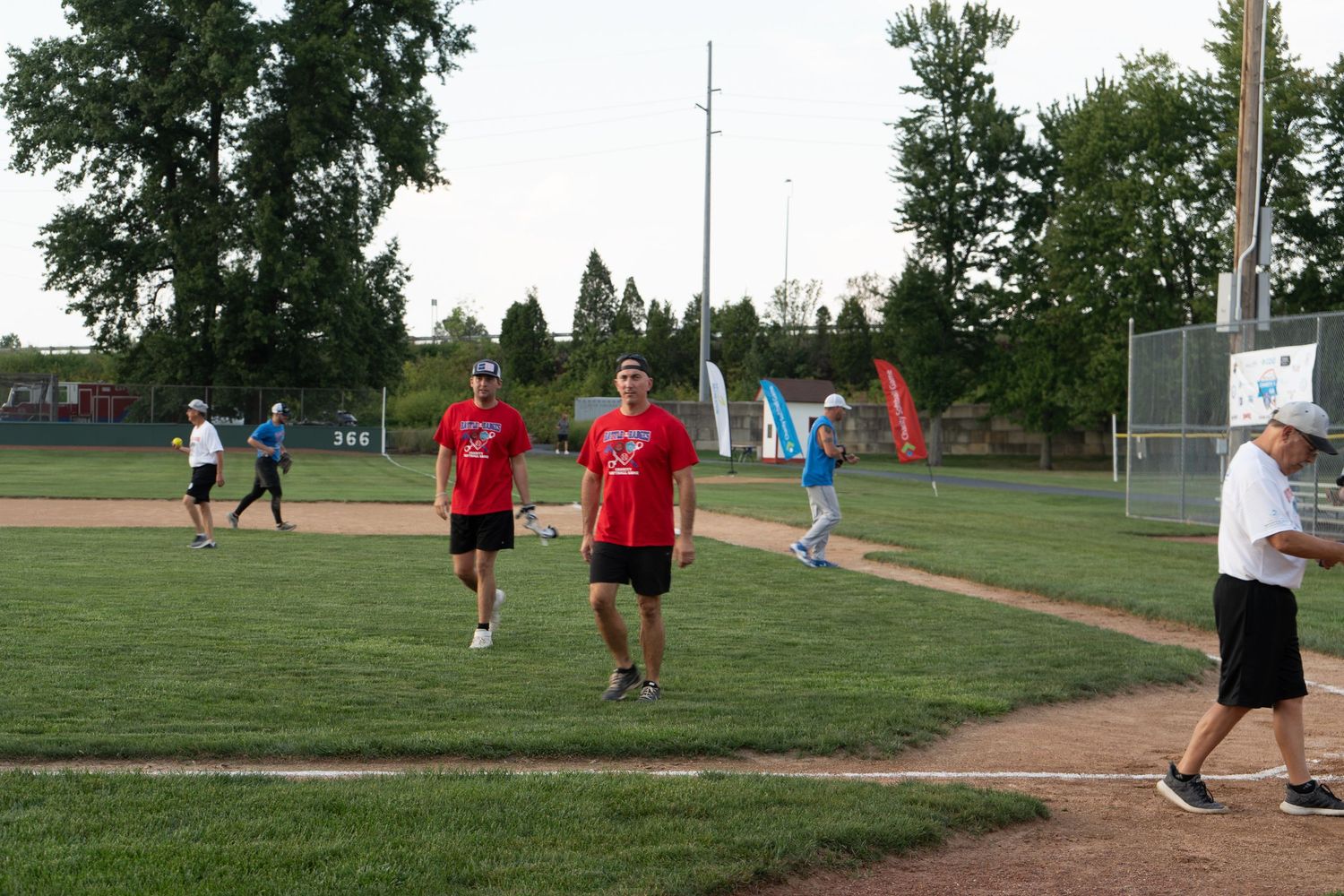 Group of adults playing recreational softball on a green field during a sunny evening game.