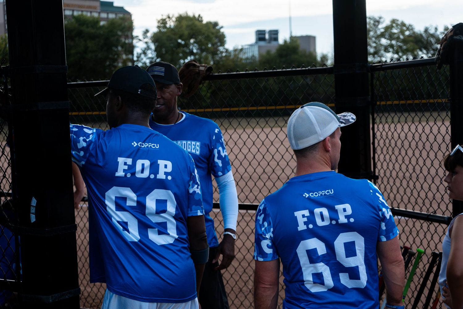 Two players in blue F.O.P. 69 softball jerseys stand together near a chain-link fence dugout.