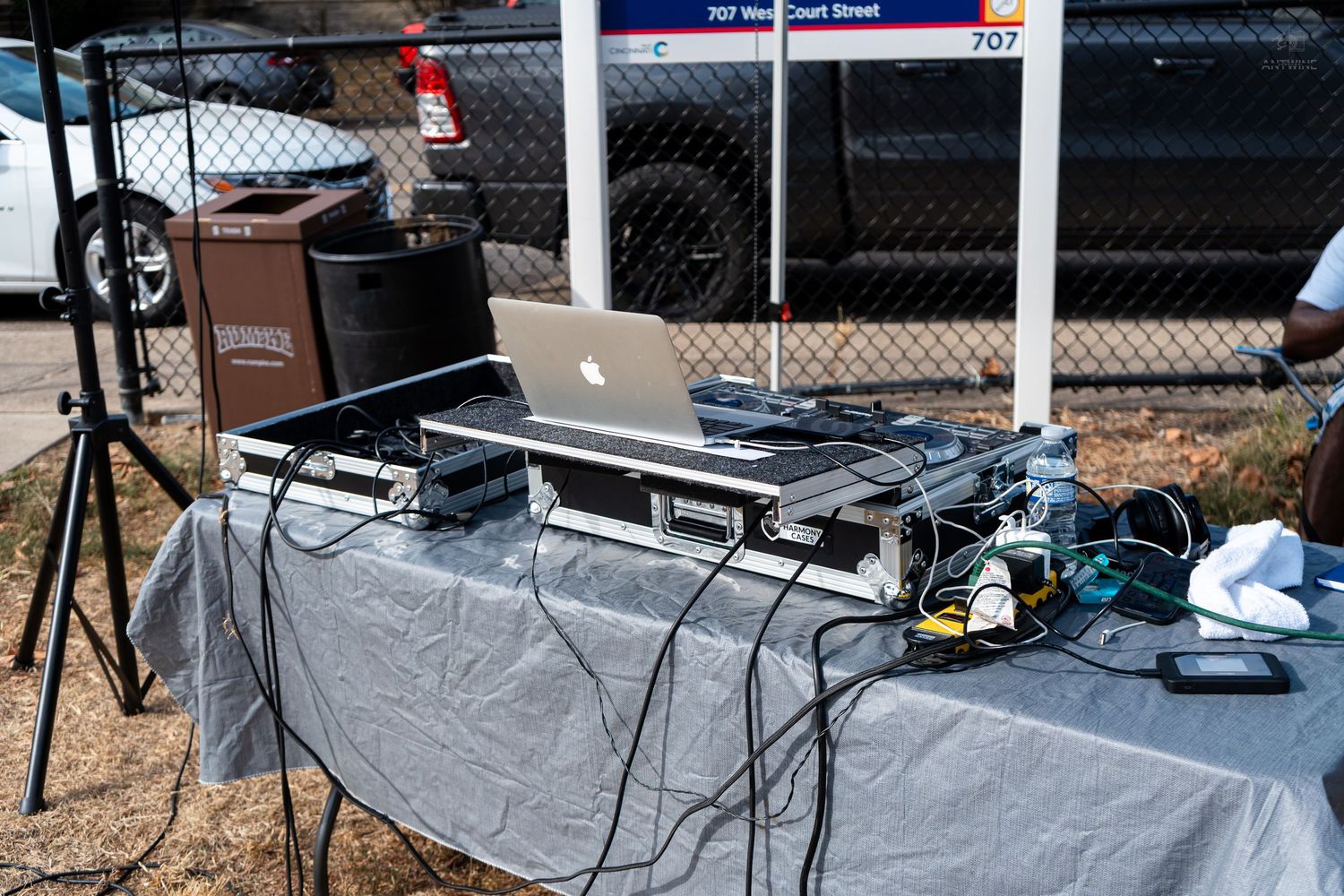 Outdoor DJ setup with laptop, mixer, and speakers on a gray draped table at a sunny event.