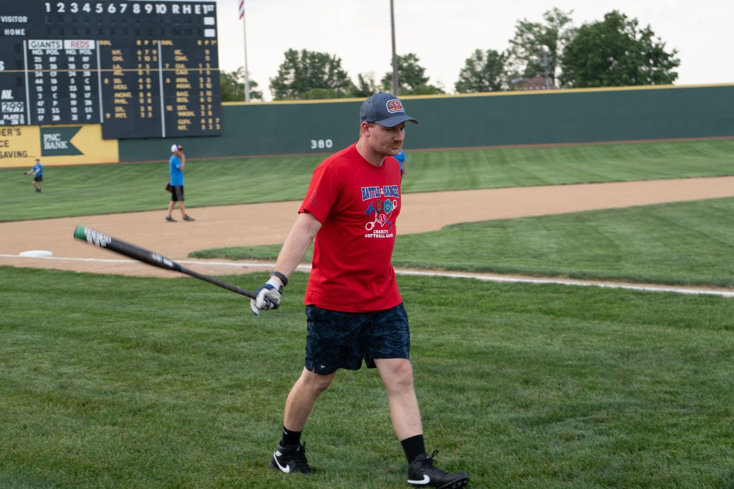 Man in red shirt holding baseball bat on green field near outfield wall during outdoor batting practice event.