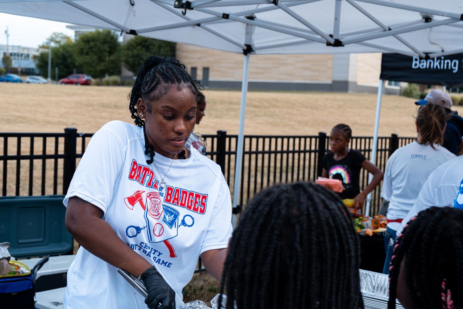 Woman in a white graphic t-shirt serving food at an outdoor community event under a canopy tent.