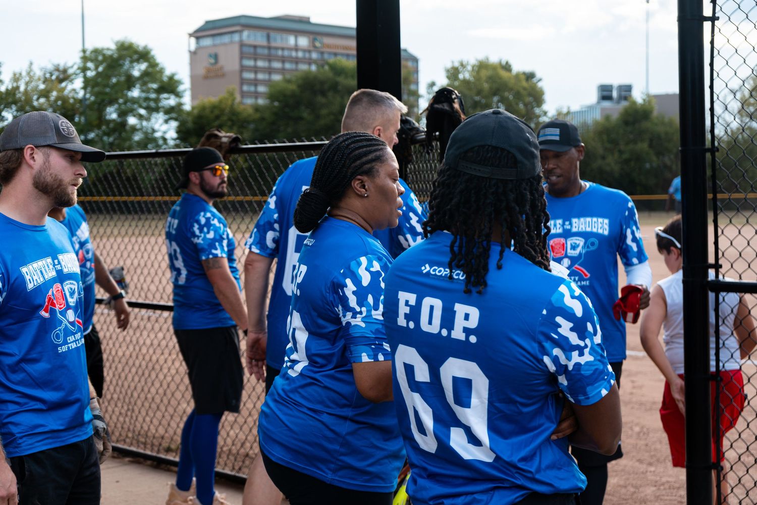 Players in blue Battle of Badges F.O.P. jerseys walk together near a fence at a softball event.