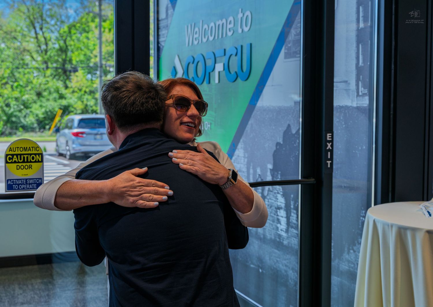 Two people share a warm hug inside a COPFCU credit union branch during a welcoming event.