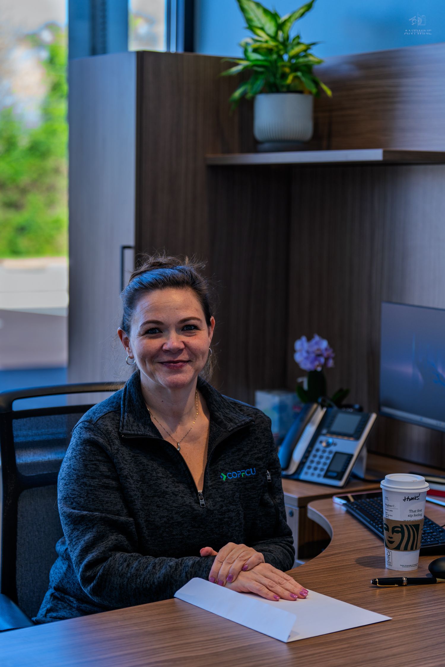 A smiling woman in a dark fleece jacket sits at a modern office desk with a Starbucks coffee cup and documents.