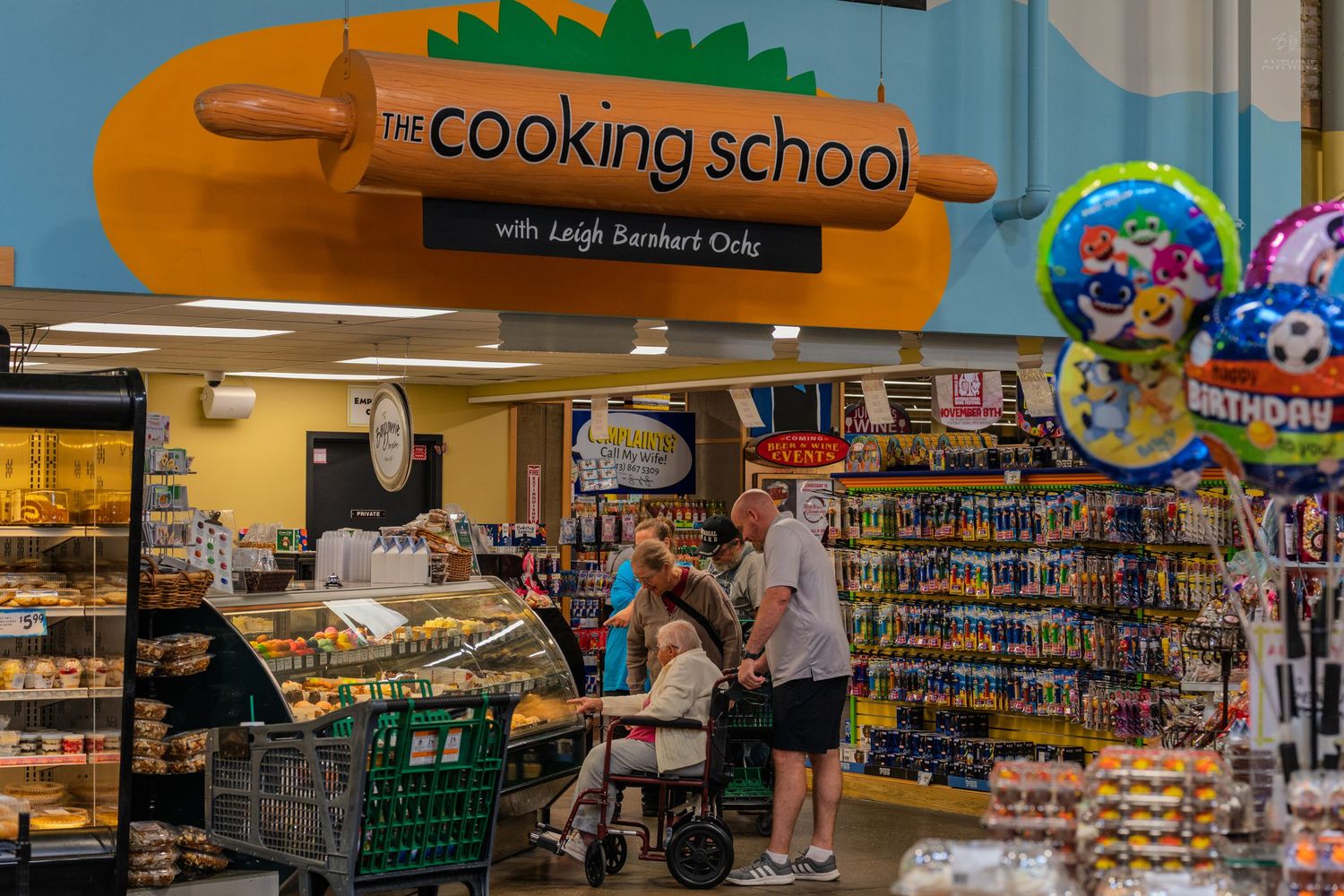 The Cooking School with Leigh Barnard Ochs sign above a deli counter in a grocery store.