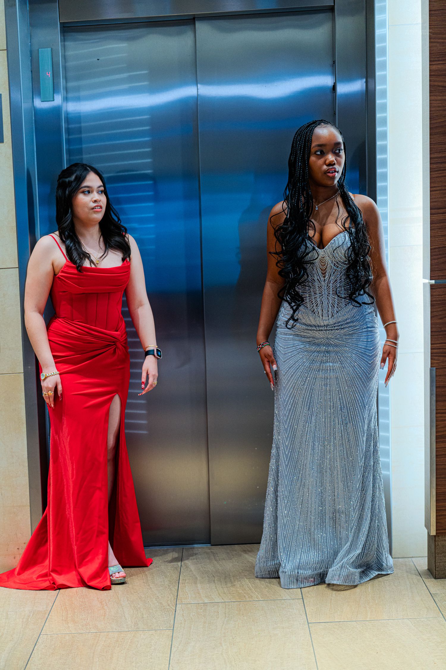 Two young women in formal red and silver mermaid gowns posing elegantly in front of a metallic elevator.