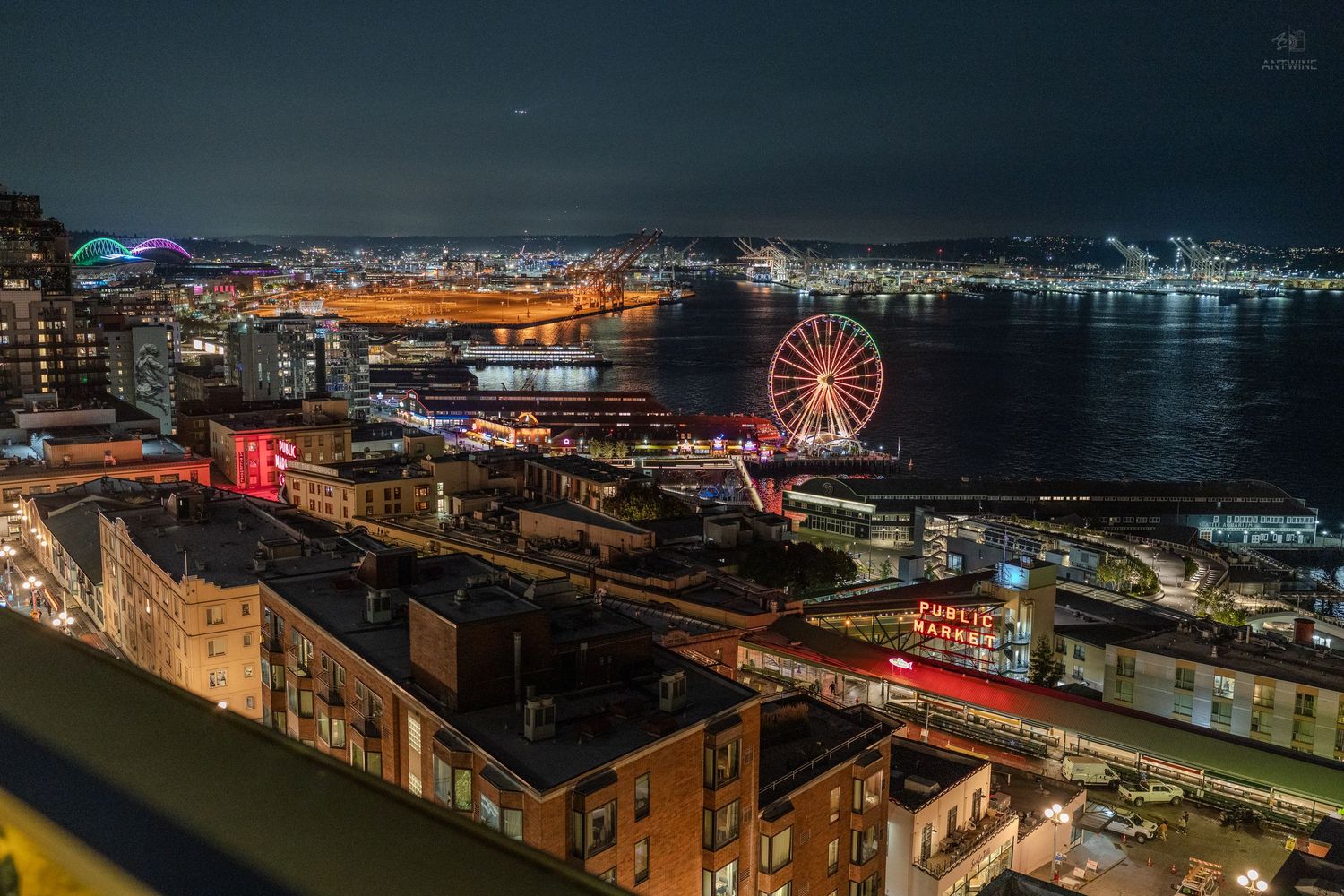 Aerial night view of Seattle waterfront featuring the Great Wheel and city lights reflecting on water.