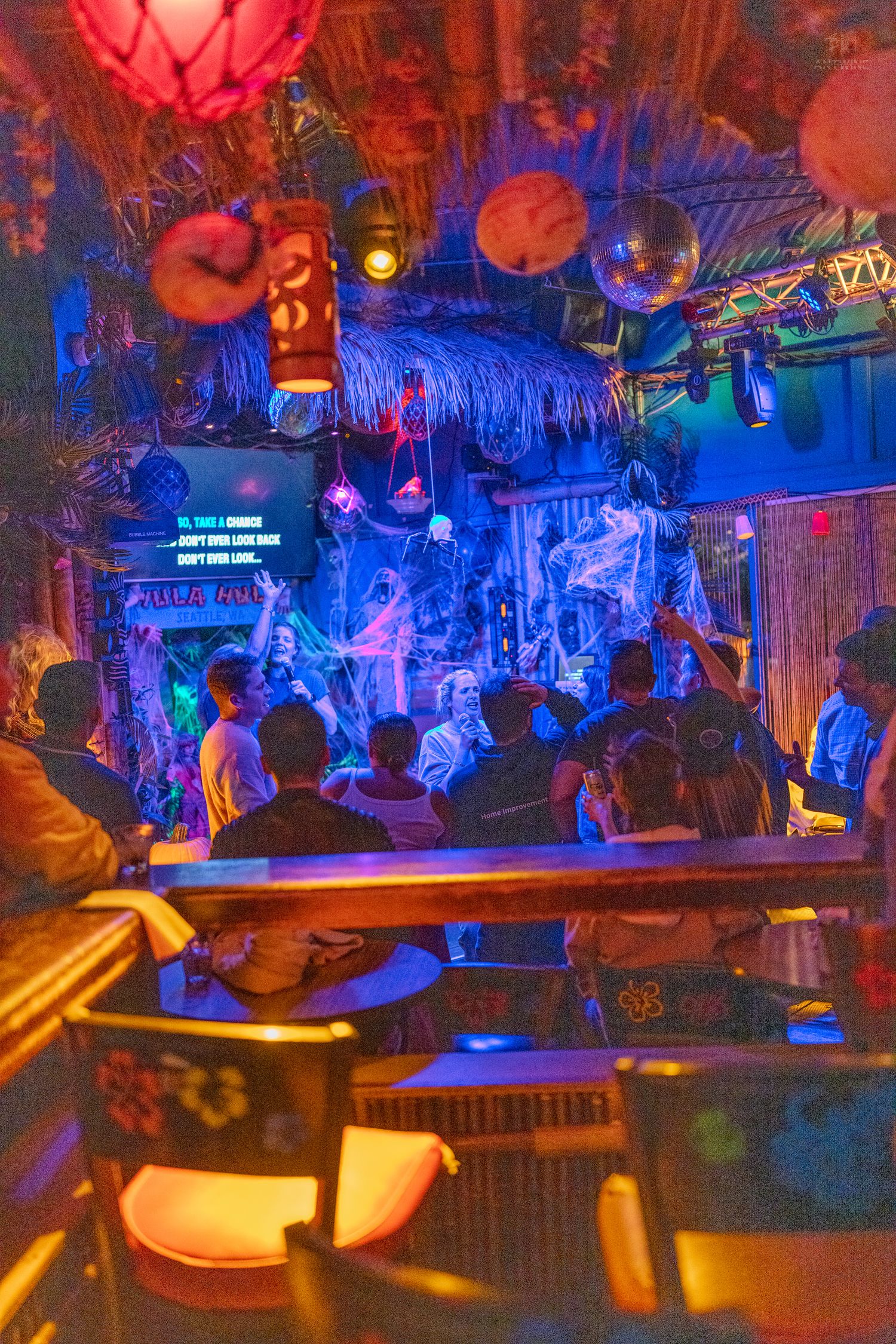 Colorful tiki bar interior with neon lights, tropical decor, and guests watching a themed stage performance.