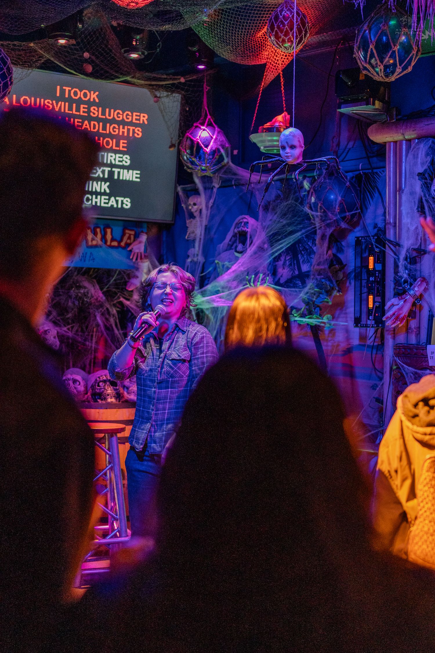 Woman singing karaoke at spooky Halloween-themed Hula Hula bar with cobweb decorations.