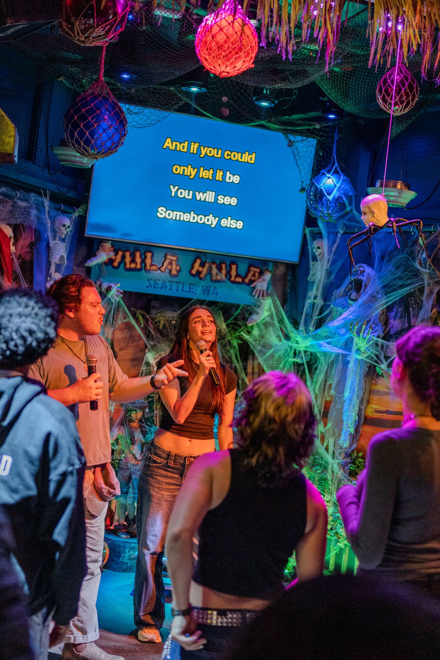 Couple singing Somebody Else karaoke at colorful Halloween-decorated Hula Hula bar.