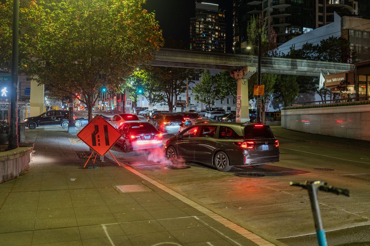 Night urban street scene with cars, orange construction barriers, and city buildings illuminated in the background.