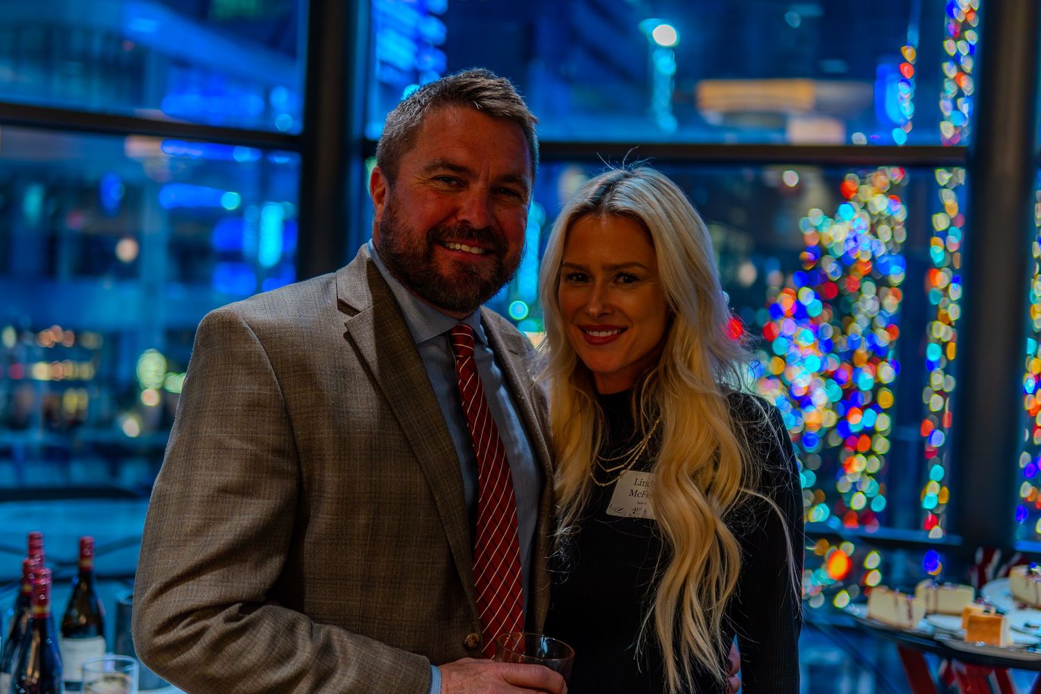 A smiling couple poses together at a festive evening event with colorful bokeh lights in the background.
