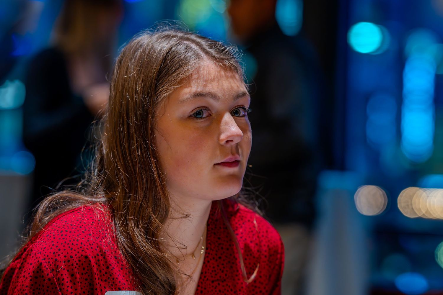 Young woman in red top with long brown hair gazing thoughtfully in a softly lit indoor setting.