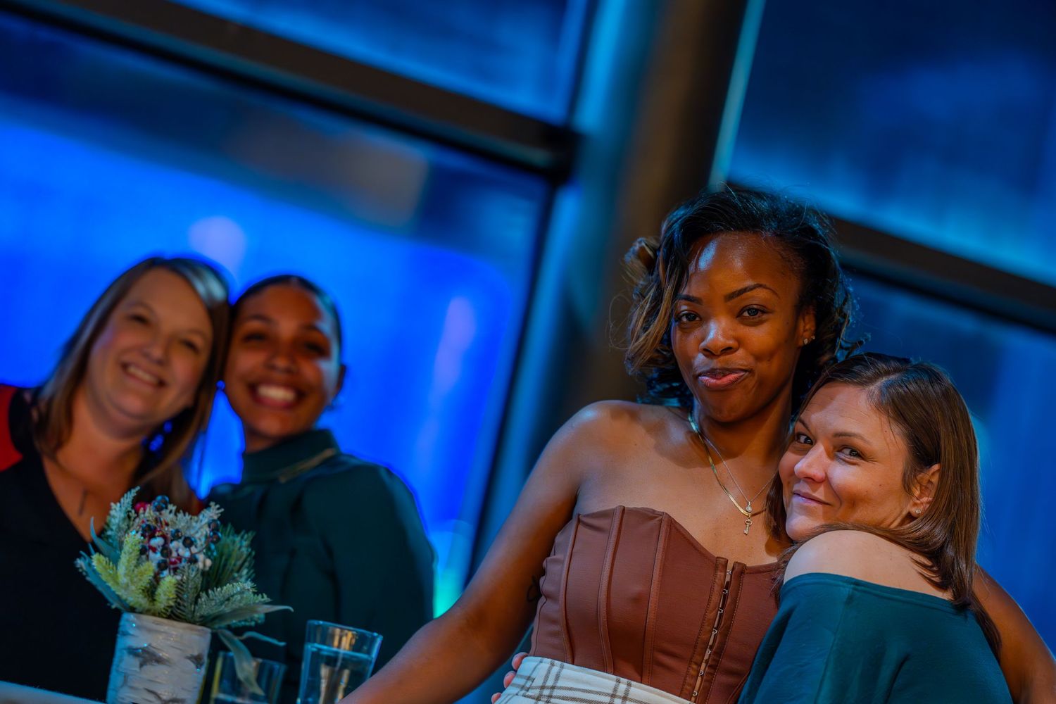 Four women smiling and posing together at an indoor event with blue ambient lighting in the background.