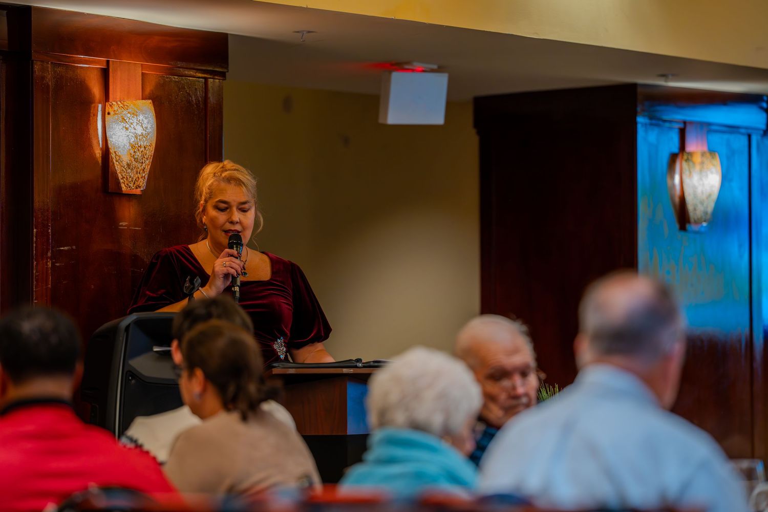 Woman speaking into microphone at indoor restaurant event with seated audience in warm ambient lighting.