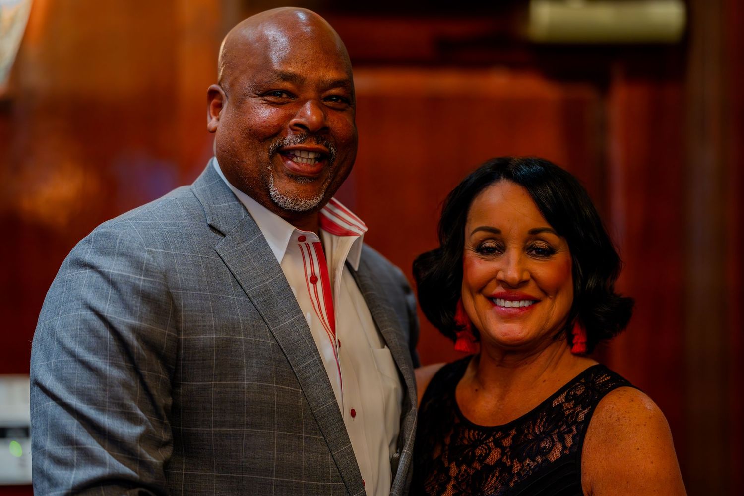 A smiling man in a gray suit and a woman in a black dress with red earrings pose together at a formal event.