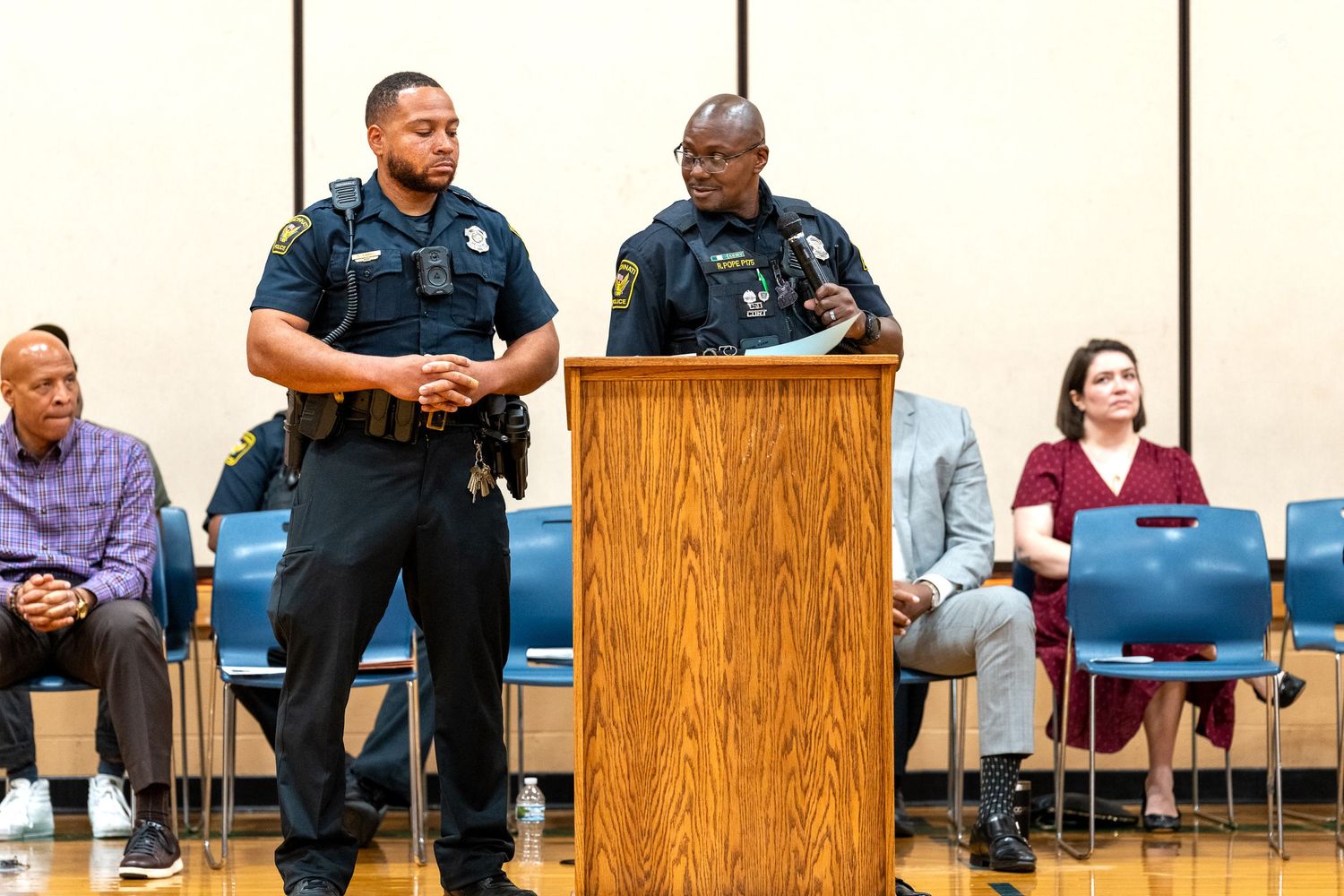 Two police officers stand at a podium addressing an audience during a community meeting in a public hall.