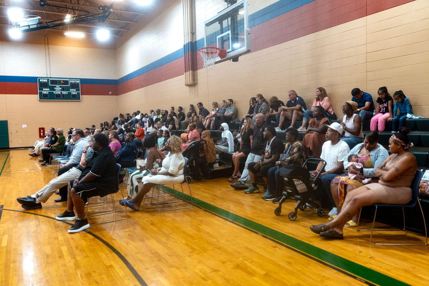 Large crowd of diverse people seated in bleachers inside a school gymnasium during a community event.