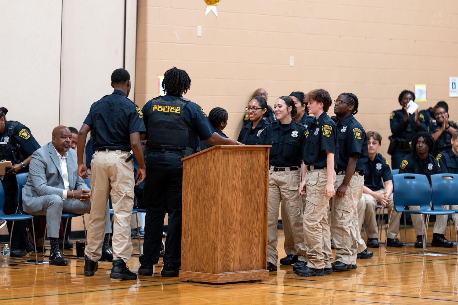 Police officers and youth cadets gather around a podium during an indoor ceremony in a school gymnasium.