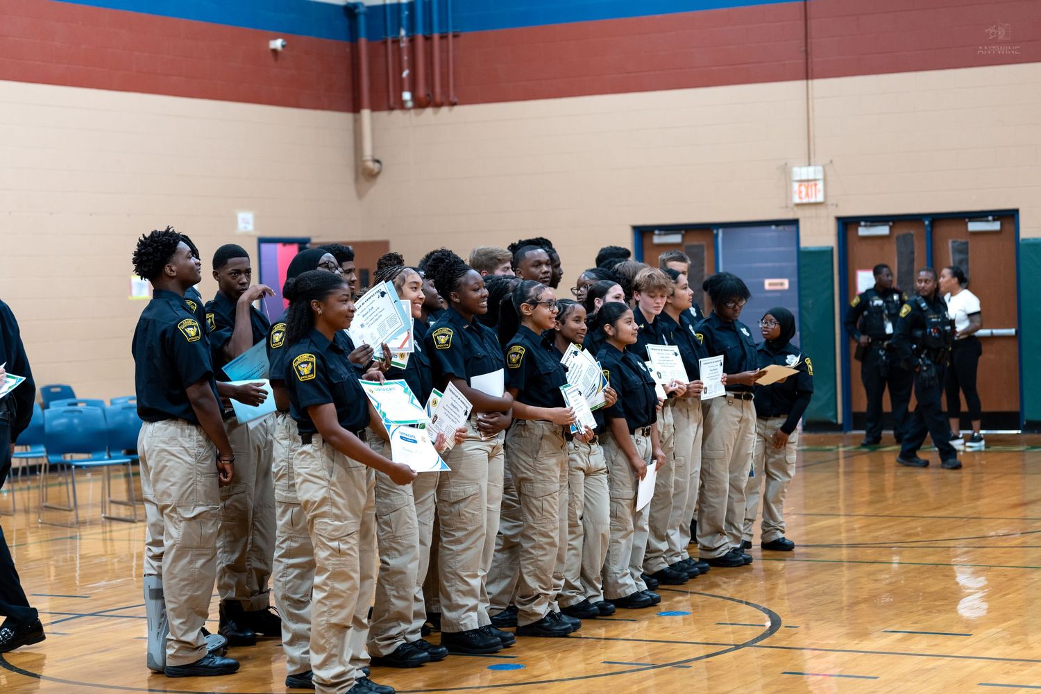 Young students in uniform shirts and khaki pants stand in a line during an awards ceremony in a school gymnasium.