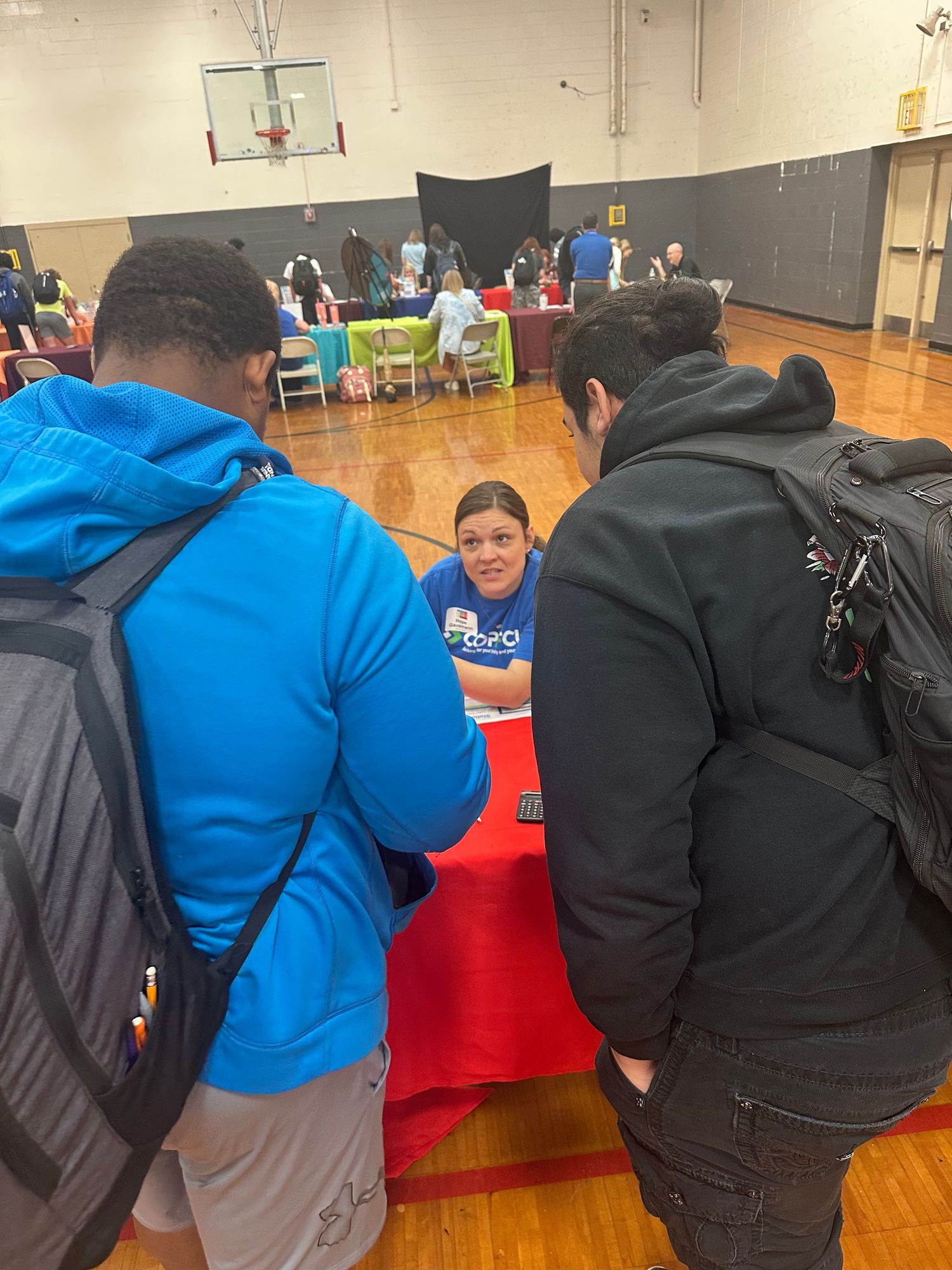 Students talk with a recruiter at a red table during an indoor career fair in a school gymnasium.