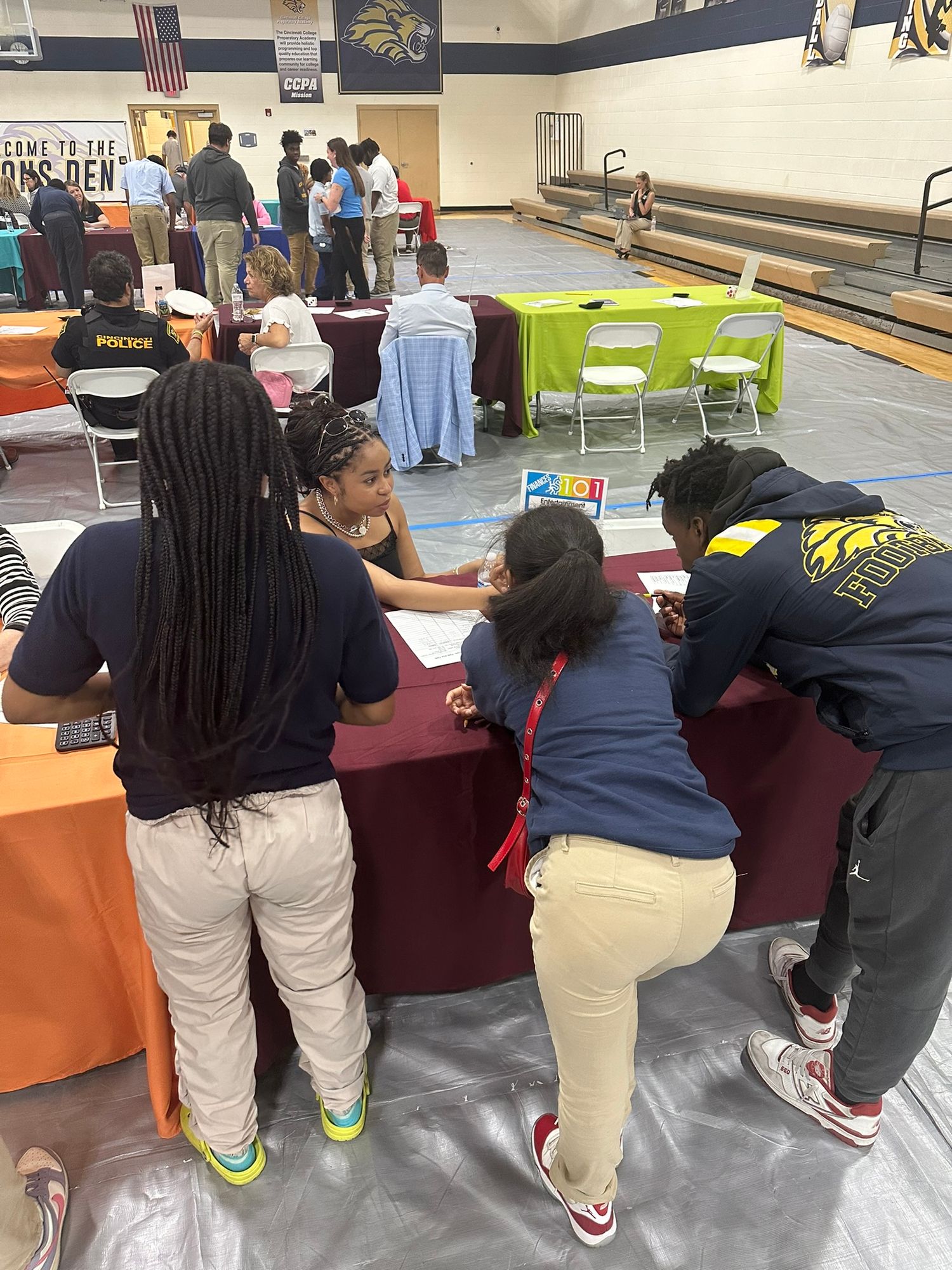 Students gather around a maroon table at a school career fair held in a gymnasium with colorful booths.