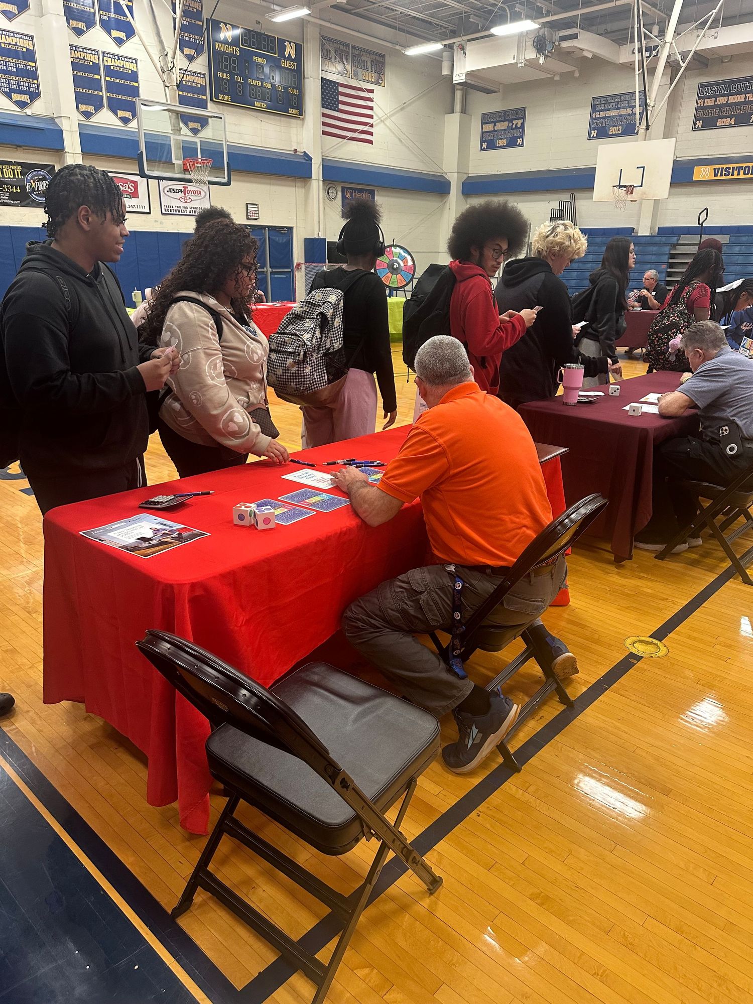 Students browse career fair tables in a school gymnasium with red-clothed tables and representatives sharing information.