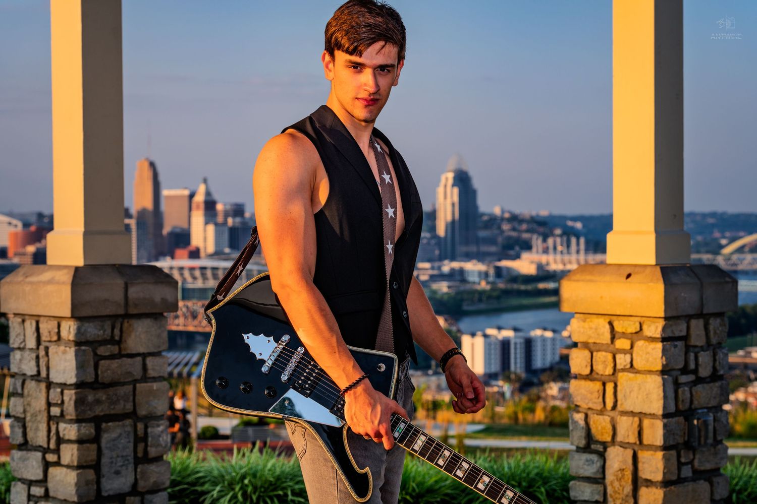 Young male guitarist holding black electric guitar with city skyline backdrop at golden hour.