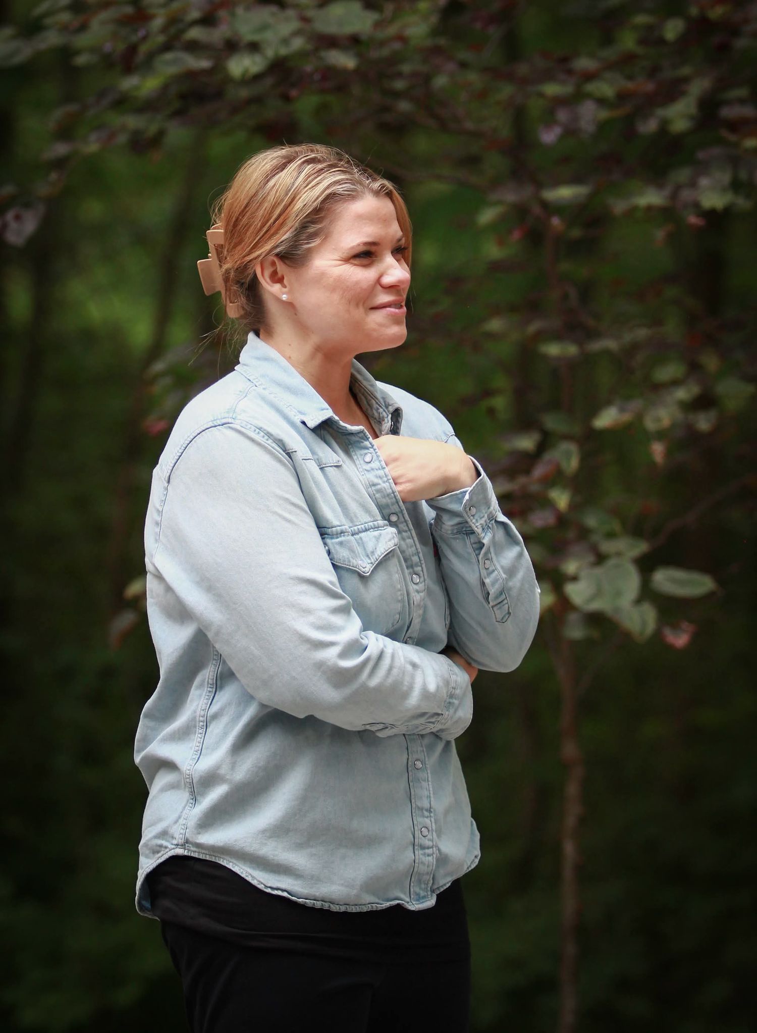 Woman in denim shirt standing outdoors with arms crossed, smiling in a wooded setting.
