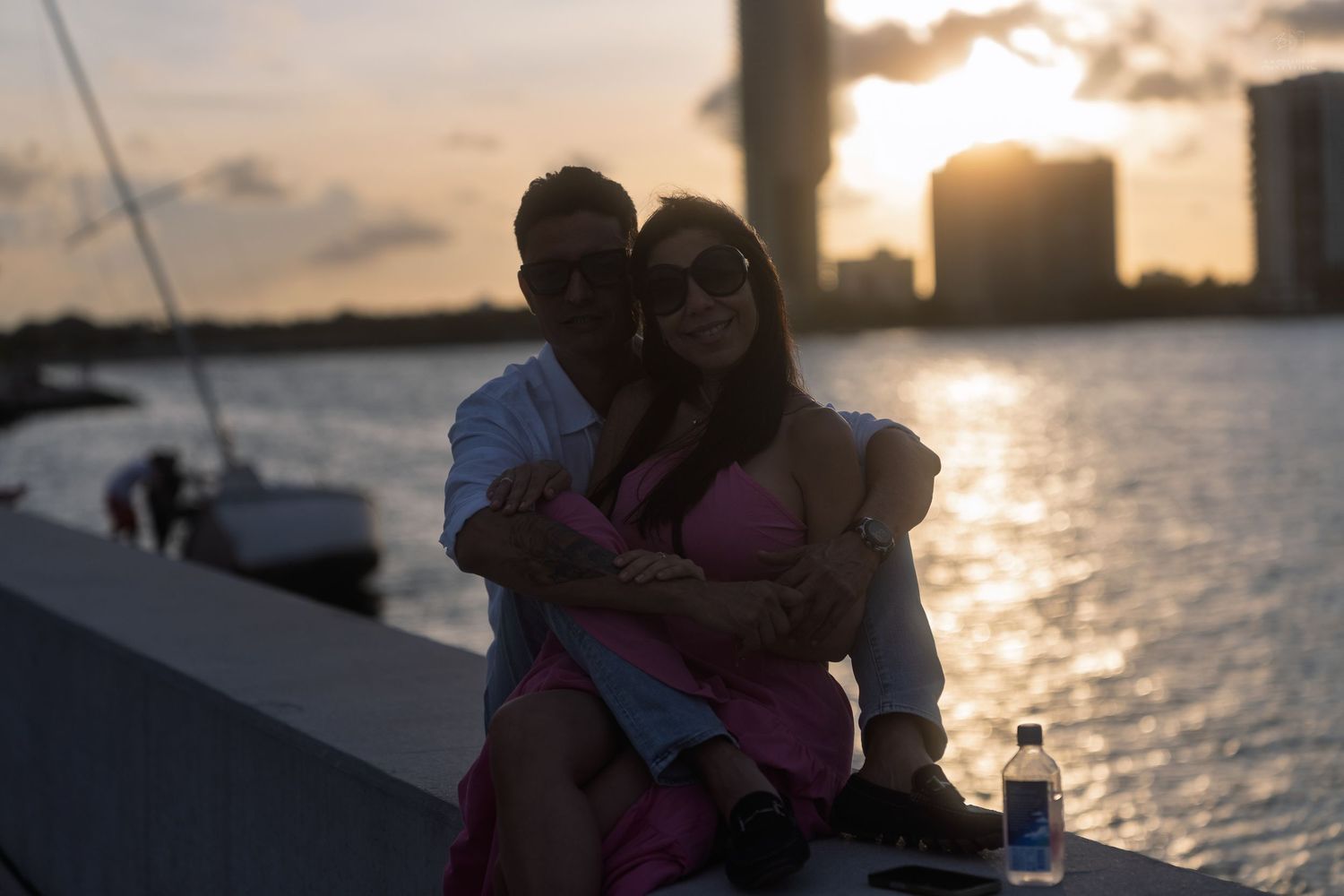 Romantic couple embracing on a waterfront wall at sunset with city skyline and sailboat in background.