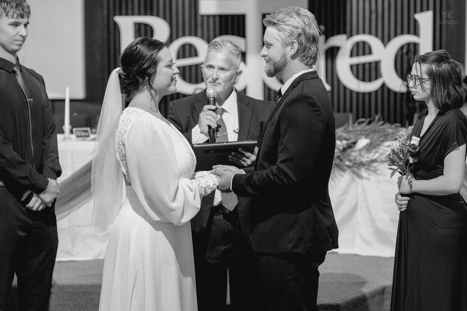 Couple holds hands at altar during indoor wedding ceremony with officiant and wedding party.