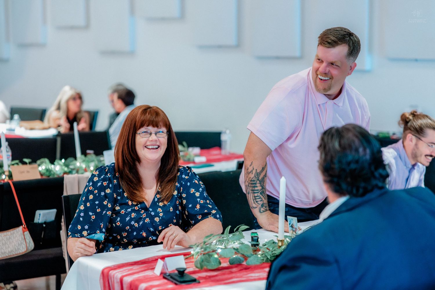 A woman with red hair laughs joyfully with a tattooed man at a wedding reception table.