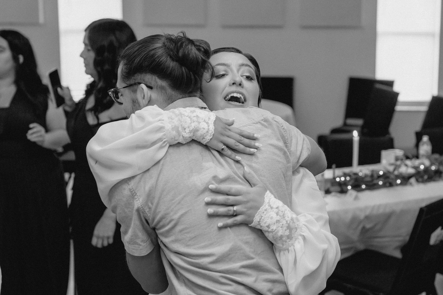 Bride laughing joyfully while embracing a guest at a wedding reception in black and white photo.