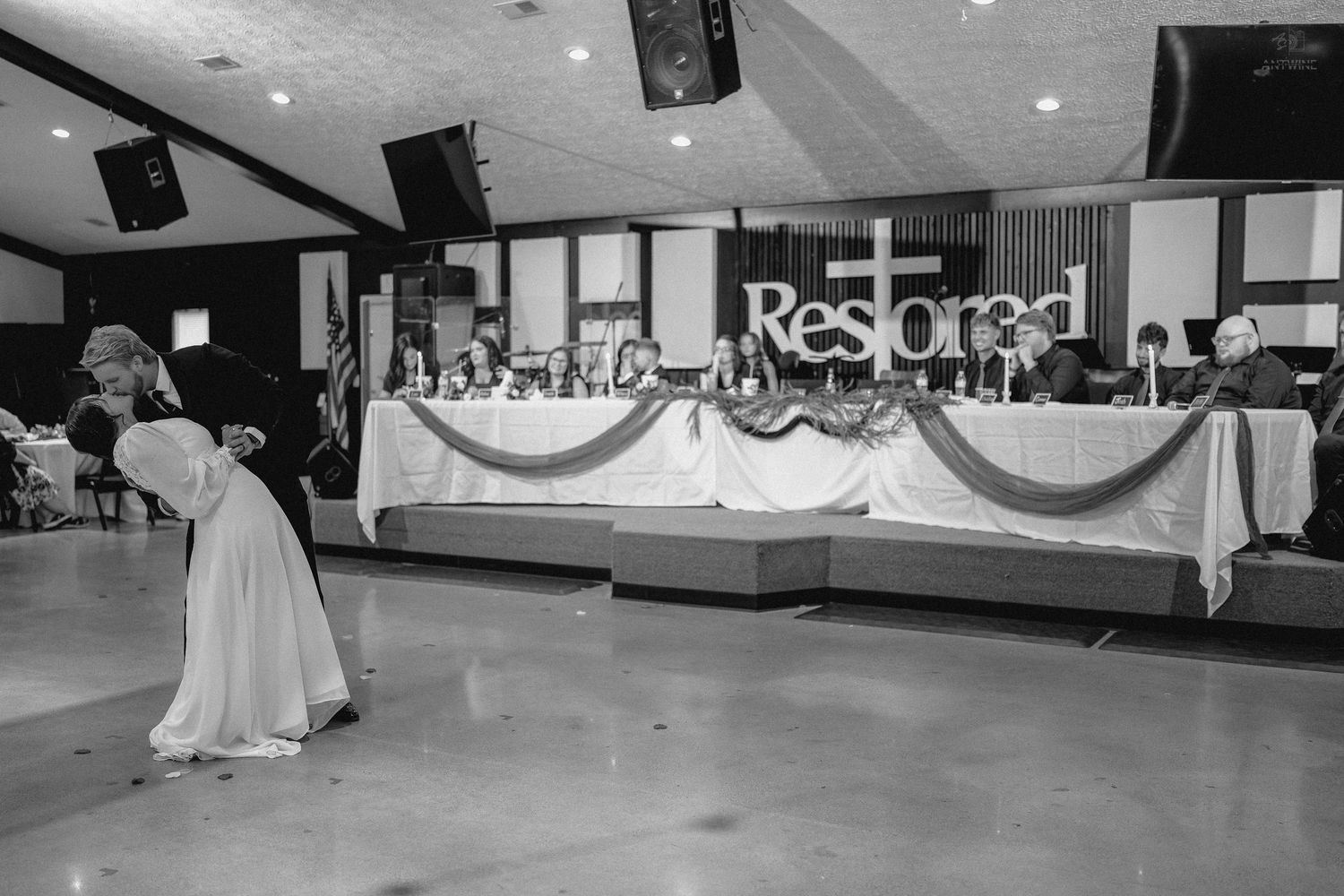 Wedding reception hall with bridal party at head table beneath a Restored sign during first dance.