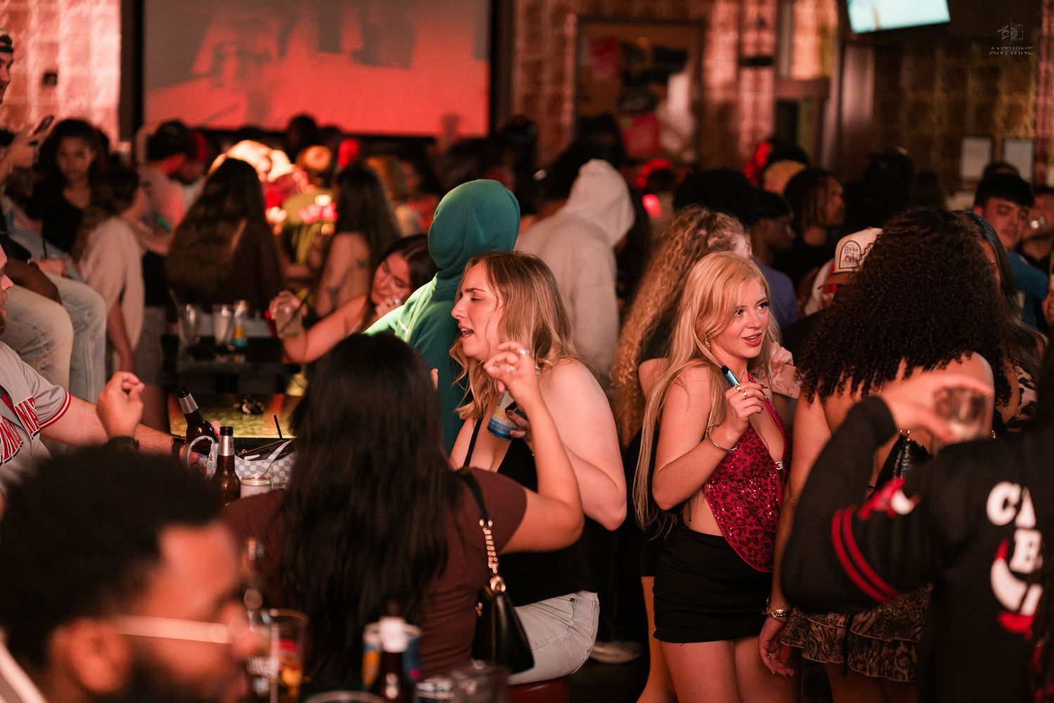 Crowded bar scene with diverse group of young adults socializing and drinking in dimly lit nightclub.